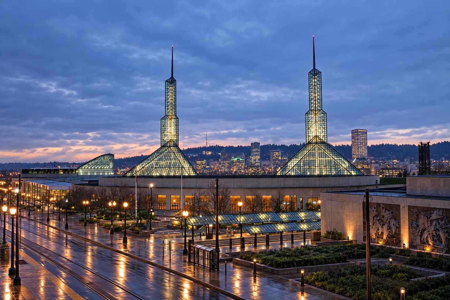 The two glass spires of the Oregon Convention Center in Portland lit at dusk, with the MAX light-rail tracks and wet plaza in the foreground