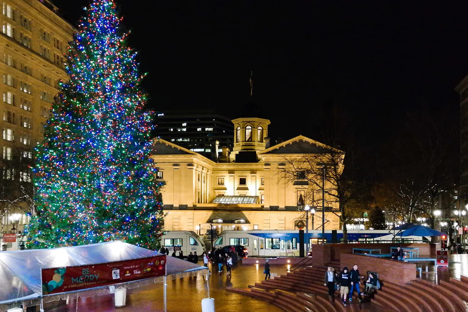The lit Christmas tree in Pioneer Courthouse Square at night, with the glowing Pioneer Courthouse behind it and a MAX train passing