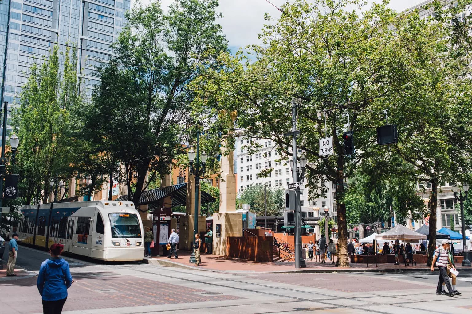 A MAX light-rail train stopped beside Pioneer Courthouse Square in downtown Portland, with tall street trees and market tents around the square.