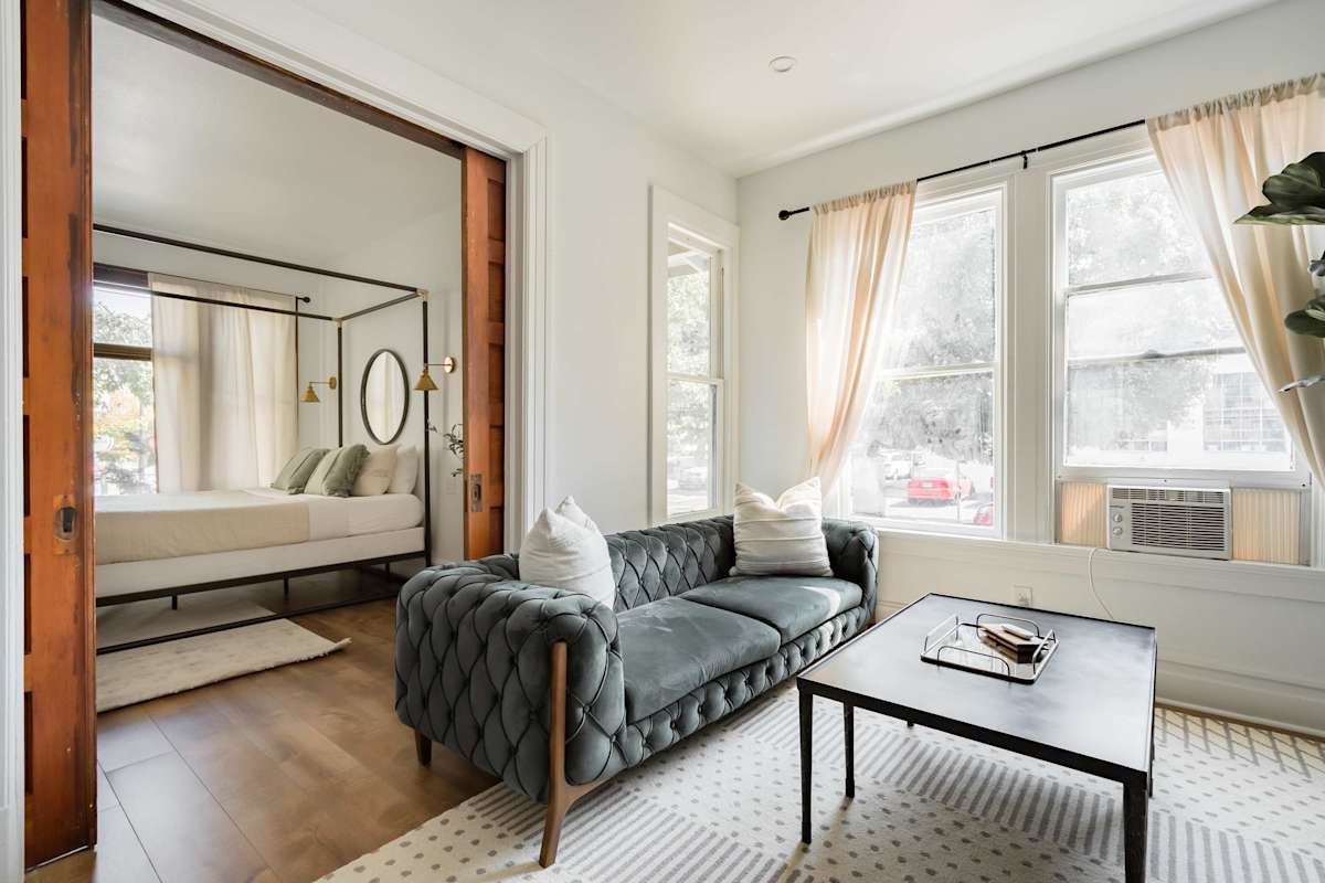 A charming apartment living room with teal velvet sofa, leather accent chair, and canopy bed visible through doorway